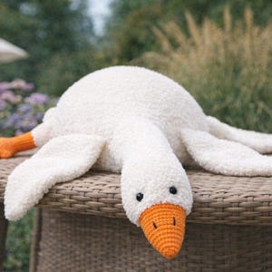 May include: A plush white goose with an orange beak and feet lies on a wicker surface. The goose has black eyes and a soft, textured body. The background features blurred greenery.