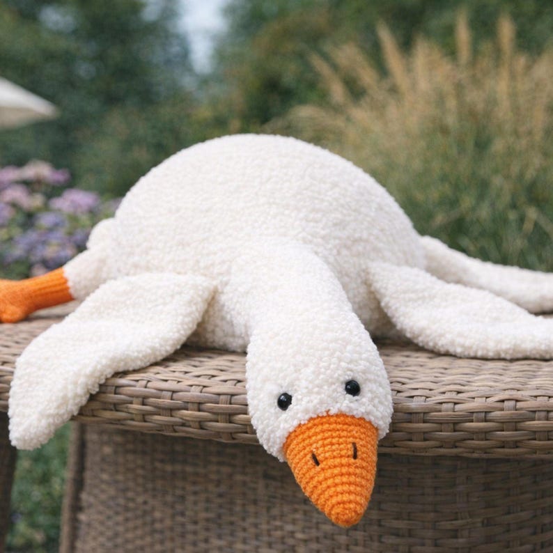 May include: A plush white goose with an orange beak and feet lies on a wicker surface. The goose has black eyes and a soft, textured body. The background features blurred greenery.