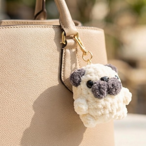 Può includere: Un portachiavi a forma di carlino in peluche color crema, con dettagli grigi sulle orecchie e sul muso. Il portachiavi è attaccato a una borsa beige con una chiusura dorata. Il carlino ha occhi neri e una consistenza soffice.