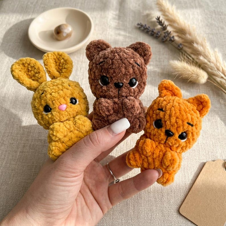 May include: Three handmade crochet stuffed animals: a yellow bunny with pink nose, a brown bear, and an orange bear. The toys are held in a hand, with a neutral background. The toys are made of soft, textured yarn.