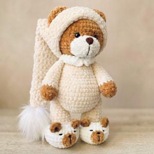 May include: A handmade crochet teddy bear wearing a cream-colored outfit with a matching hat and bear-shaped slippers. The bear has brown accents on its paws, ears, and snout. The hat has a long, cream-colored tail with a white fluffy pom-pom.