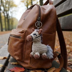 Puede incluir: Un llavero de caballo de ganchillo en beige, marrón y gris cuelga de una mochila de cuero marrón. El caballo tiene una cola gris esponjosa y cascos marrones. La mochila es de color marrón intenso y tiene cremallera y varios bolsillos.