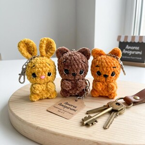 May include: Three handmade plush amigurumi keychains: a yellow bunny, a brown bear, and an orange bear. Each keychain has a metal clasp and a small tag that reads "Amoree Amigurumi Handmade Plush Friends Set of 3."