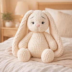 May include: A cream-colored, handmade crochet bunny with long floppy ears, a pink nose, and black stitched eyes. The plush toy is sitting on a bed with a neutral-toned blanket. The bunny is a soft, cuddly stuffed animal.