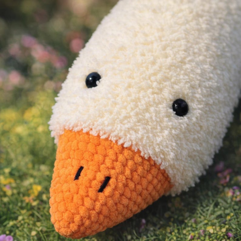 May include: Close-up of a plush white goose head with an orange beak and black bead eyes. The soft, textured fabric creates a cuddly appearance. The handmade goose is likely a decorative item or a child's toy.