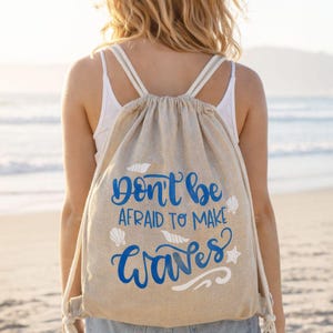 Könnte beinhalten: Beigefarbener Rucksack mit Kordelzug und dem blauen Schriftzug "Don't be afraid to make waves". Die Tasche hat weiße Träger und wird von einer Person am Strand getragen. Die Tasche hat Muschel- und Sterngrafiken.