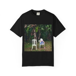 Op de afbeelding: Zwart t-shirt met een afbeelding van een cartoonkonijn dat in een tropische omgeving op een witte stoel zit. Het konijn draagt een overall en houdt een drankje vast. Een tweede witte stoel is leeg. Groene bananenbomen op de achtergrond.