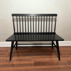 May include: A black wooden bench featuring a spindle backrest with eleven vertical slats, a flat rectangular seat, and four tapered legs connected by horizontal support stretchers. The bench has a smooth finish and a logo reading NORRGRAIN.
