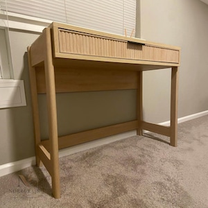 May include: A light-toned wooden desk featuring a single drawer with a vertically fluted, textured front panel. The desk is supported by four straight legs connected by a horizontal support beam, showcasing a natural wood grain finish throughout.
