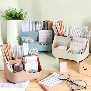 May include: Three pastel-colored desk organizers filled with pencils, pens, sticky notes, and other office supplies. The organizers are on a wooden desk, with a small plant in the background. A pair of glasses and a pen are also on the desk.