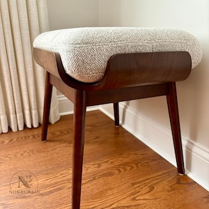 May include: A rectangular ottoman featuring a dark brown curved wooden frame and four tapered legs. The top is upholstered in a textured, light beige fabric with a subtle grid pattern. The piece is marked with the Norrgrain logo on the lower left.