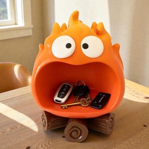 May include: Orange, cartoon-style storage container shaped like a flame with large white-rimmed eyes. The container holds keys and a remote. It rests on two brown log-shaped supports, adding a whimsical touch to home decor.