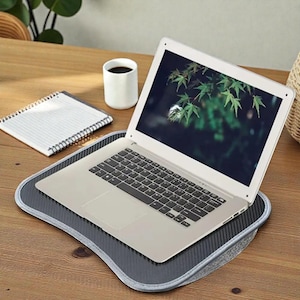 May include: A silver laptop rests on a gray and black laptop desk. The laptop screen displays a nature scene with green leaves. A white coffee mug and a spiral notebook are also on the wooden surface.