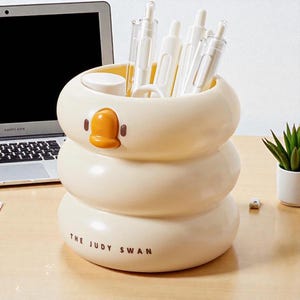 May include: A cream-colored desk organizer shaped like a swan, with a yellow beak and the text "THE JUDY SWAN". The organizer holds several white pens and pencils. A small succulent plant and a laptop are visible in the background.