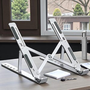 May include: Silver laptop stand with black rubber pads, designed to elevate a laptop. The stand is on a wooden desk, with a notebook, pen, and keys nearby. The stand is adjustable and foldable.
