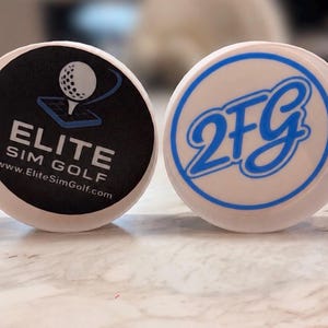May include: Two circular white plastic caps. The left cap features a black circular insert with a white golf ball graphic, the text ELITE SIM GOLF, and the website www.EliteSimGolf.com. The right cap has a white insert with the blue text 2FG.