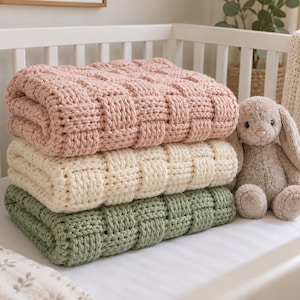 May include: A stack of three chunky knit blankets in soft pastel colors including dusty pink, cream, and sage green, featuring a textured basketweave pattern. Beside the folded blankets sits a plush, light brown stuffed rabbit toy.