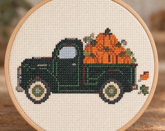 May include: A cross-stitch embroidery in a round wooden hoop featuring a dark green vintage pickup truck carrying a large pile of bright orange pumpkins on a cream-coloured fabric background. The truck has light blue windows and green wheels.