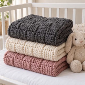 May include: A stack of three chunky knit blankets in dark gray, cream, and dusty rose colors, featuring a textured basketweave pattern. A plush beige stuffed rabbit toy sits beside the folded blankets on a white surface inside a crib.