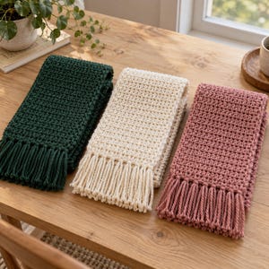 May include: Three rectangular scarves featuring a textured crochet stitch pattern. The scarves are arranged side-by-side in dark forest green, cream, and dusty rose pink. Each scarf is finished with a row of matching fringe along the bottom edge.