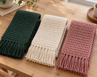 May include: Three rectangular scarves featuring a textured crochet stitch pattern. The scarves are arranged side-by-side in dark forest green, cream, and dusty rose pink. Each scarf is finished with a row of matching fringe along the bottom edge.