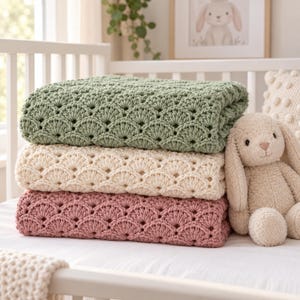 Può includere: Una pila di tre coperte all'uncinetto nei colori verde salvia, crema e rosa antico, ciascuna caratterizzata da un motivo a ventaglio ripetuto. Un peluche a forma di coniglio beige è seduto accanto alle coperte piegate su una superficie bianca.