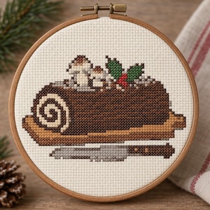 May include: A circular wooden embroidery hoop containing a cross-stitch pattern of a chocolate yule log cake. The cake features a spiral end, light brown base, and is decorated with small mushrooms, green holly leaves, and red berries, with a knife below.