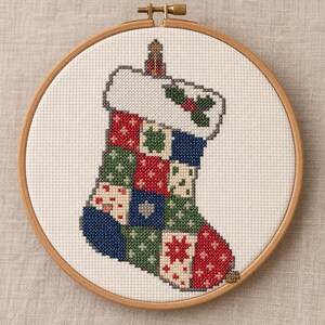 Può includere: Un ricamo a punto croce di una calza natalizia patchwork in un telaio di legno rotondo. La calza presenta un risvolto bianco con foglie di agrifoglio e bacche rosse, e un corpo composto da quadrati rossi, verdi, blu e bianchi con motivi.
