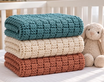 May include: A stack of three chunky knit blankets in teal, cream, and terracotta colours, featuring a textured basketweave pattern. Beside the folded blankets sits a plush, light beige stuffed rabbit toy with long ears and a soft, fuzzy texture.