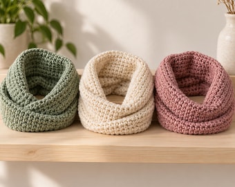 May include: Three circular infinity scarves arranged in a row, featuring a chunky crochet knit texture. The scarves are coloured in sage green, cream, and dusty rose. Each piece is constructed with a consistent, thick yarn loop design.
