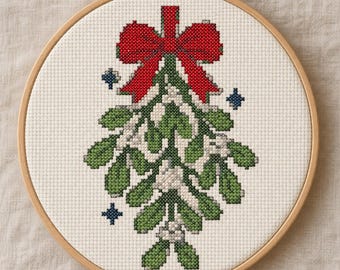 May include: A cross-stitch embroidery piece in a round wooden hoop featuring a design of green mistletoe leaves and white berries tied with a large red bow. Small dark blue star-like accents are positioned on either side of the mistletoe.