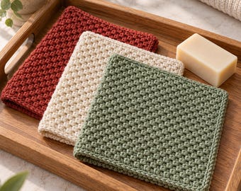 May include: Three square, hand-crocheted washcloths in solid dark red, cream, and sage green colours. Each cloth features a dense, textured, raised stitch pattern and a finished border. A rectangular bar of light beige soap sits beside them.