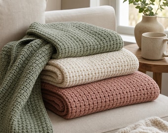 May include: A stack of three folded blankets featuring a textured waffle knit pattern. The top blanket is sage green, the middle is cream, and the bottom is a muted terracotta colour. The blankets are draped over a light-coloured upholstered sofa.