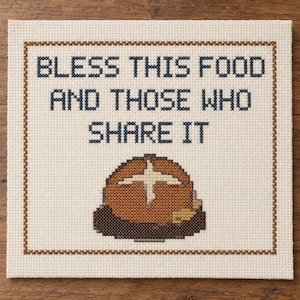 May include: A square cross-stitch embroidery on cream-coloured fabric featuring a brown bread loaf with a cross pattern. The text reads: BLESS THIS FOOD AND THOSE WHO SHARE IT in dark blue letters, enclosed by a brown stitched border frame.