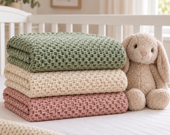May include: A stack of three folded knit blankets in sage green, cream, and dusty rose colours, each featuring a textured waffle stitch pattern. Beside the blankets sits a plush, light beige stuffed rabbit toy with long ears and dark eyes.