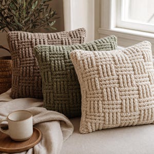 May include: Three square throw pillows featuring a chunky basketweave knit pattern. The pillows are arranged in a row, colored in shades of light brown, sage green, and cream. A ceramic mug sits on a small wooden coaster in the foreground.