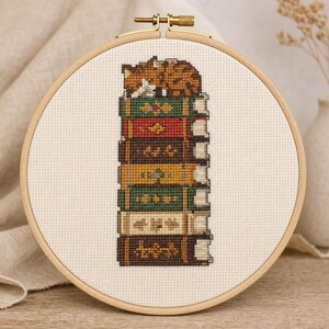 May include: A cross-stitch embroidery in a round wooden hoop featuring a stack of seven colorful books with decorative spines. A small orange tabby cat is curled up and sleeping on top of the highest book. The design is stitched on white fabric.