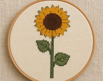 May include: A cross-stitch embroidery of a single sunflower with a dark brown centre and bright yellow petals, featuring a green stem and two green leaves, displayed within a circular wooden embroidery hoop on a light beige fabric background.