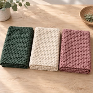 May include: Three rectangular folded cloths with a textured basketweave pattern. The cloths are arranged side by side in dark forest green, cream, and dusty rose colors. Each piece features a consistent woven stitch throughout the surface.