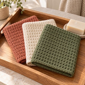 Pode incluir: Três panos de lavagem quadrados feitos à mão em crochê nas cores terracota, creme e verde-oliva. Cada pano apresenta um padrão de pontos texturizado e consistente, dobrado de forma organizada. Um sabonete retangular repousa numa bandeja.