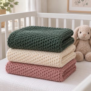 May include: A stack of three folded crochet blankets in dark green, cream, and dusty rose colors. Each blanket features a uniform, textured stitch pattern. A plush beige stuffed rabbit toy sits to the right of the stacked blankets.