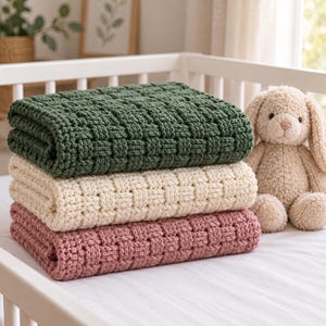 Pode incluir: Uma pilha de três cobertores dobrados com uma textura de tricô trançado grosso nas cores verde-escuro, creme e rosa antigo, posicionada ao lado de um coelho de pelúcia bege com orelhas longas e nariz bordado sobre uma superfície branca.