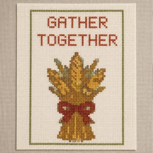 May include: A cross-stitch pattern on off-white fabric featuring the text GATHER TOGETHER in red. Below the text is a golden-yellow wheat sheaf tied with a red bow, all enclosed within a thin, dark green rectangular border stitched in a cross pattern.