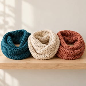 May include: Three chunky knit infinity scarves arranged in a row. The scarf on the left is deep teal, the center scarf is cream, and the scarf on the right is a muted terracotta orange. Each scarf features a consistent, textured garter stitch pattern.