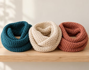 May include: Three chunky knit infinity scarves arranged in a row. The scarf on the left is deep teal, the centre scarf is cream, and the scarf on the right is a muted terracotta orange. Each scarf features a consistent, textured garter stitch pattern.