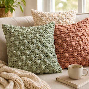 May include: Three square throw pillows featuring a textured bobble stitch pattern in sage green, terracotta orange, and cream. The pillows are arranged together, with a cream-colored knit throw blanket, a ceramic mug, and a book placed nearby.