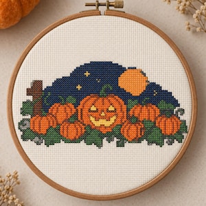 Puede incluir: Un bordado de punto de cruz en un bastidor de madera redondo que presenta una calabaza central con cara, rodeada de calabazas naranjas más pequeñas y follaje verde sobre un cielo nocturno azul oscuro con una luna y estrellas amarillas.