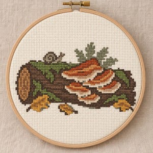 Puede incluir: Un bastidor de bordado circular de madera que contiene un diseño de punto de cruz de un tronco marrón con hongos naranjas, musgo verde, un pequeño caracol gris y hojas de otoño amarillas sobre un fondo de tela blanca.