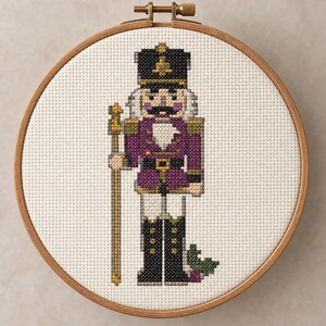 May include: A cross-stitch embroidery of a nutcracker figure in a circular wooden hoop. The nutcracker wears a black hat, a purple jacket with gold accents, and black boots, while holding a gold staff. Small green holly leaves are at the base.