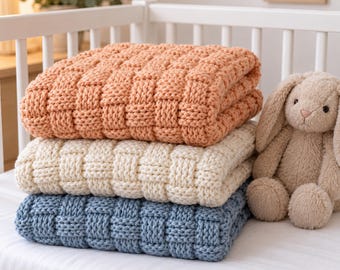May include: A stack of three chunky knit blankets in peach, cream, and dusty blue, featuring a basketweave texture. Beside the folded blankets sits a plush, light brown stuffed rabbit toy with long ears, all resting on a white surface.